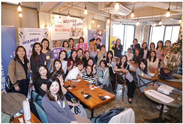 一群女性參與者聚集在溫馨明亮的咖啡廳中，進行「Women Power 台北市女性創業培訓」交流活動。參與者們面帶微笑、手持鼓勵標語如「勇敢創業」、「挺妳」等，展現積極正向氛圍。現場設有活動布條與橫幅，座位間擺有飲品與筆記用品，呈現輕鬆互動的創業社群聚會情景。