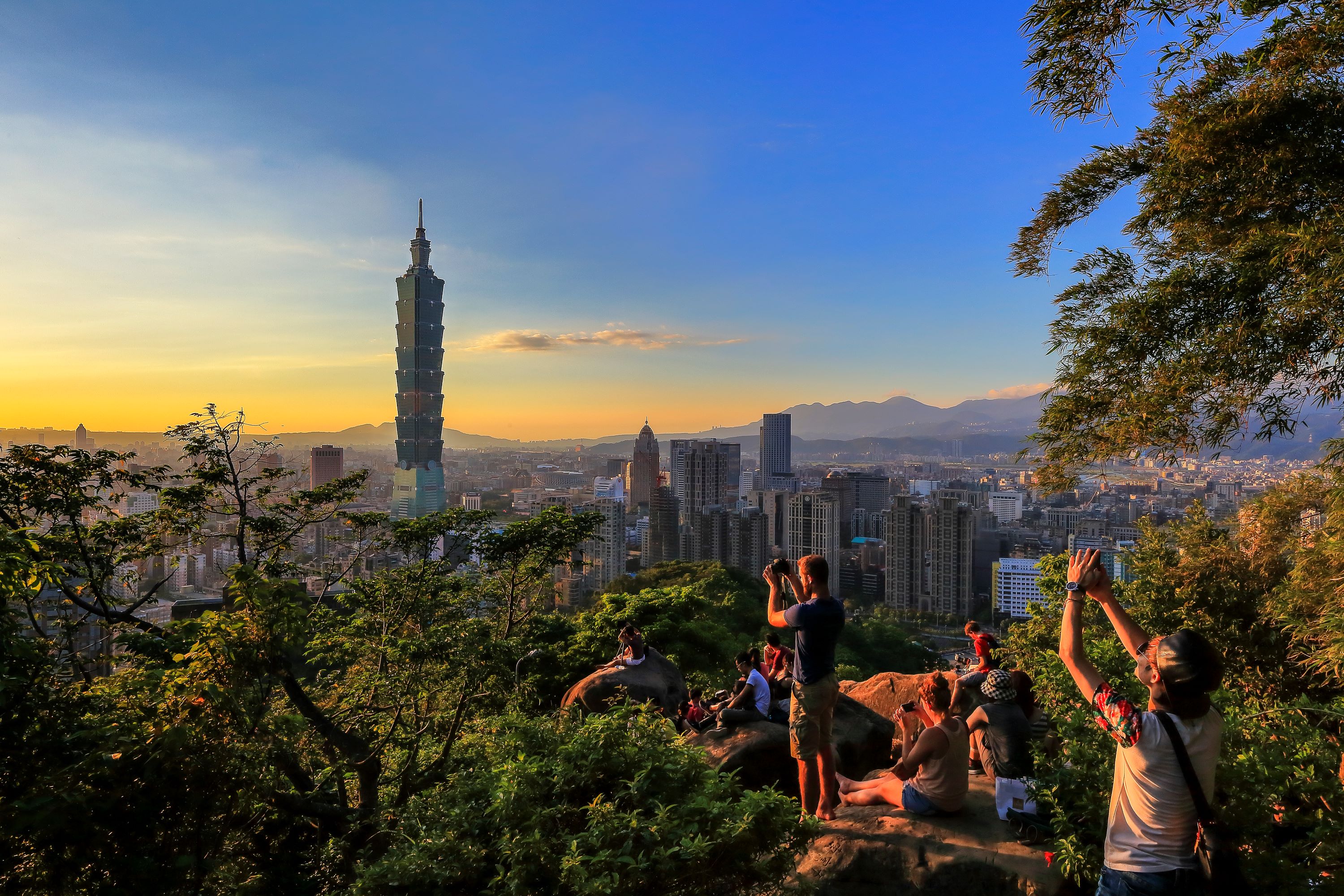 日落時分從象山眺望台北市景，畫面中央為台北101大樓，背景為落日餘暉映照的城市天際線與遠山。前景有多位遊客站立或坐在巨石上拍照與觀景，周圍被綠樹環繞，呈現自然與城市交融的景致。