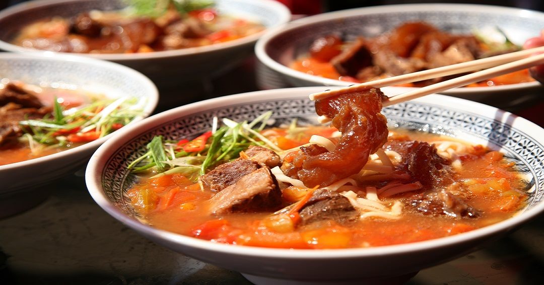 一碗熱騰騰的紅燒牛肉麵，湯頭鮮紅濃郁，內有牛肉塊、牛筋、手工麵條和蔬菜配料，畫面中筷子正夾起一塊晶亮牛筋，背景還有數碗相同牛肉麵，呈現豐盛台式美食場景。