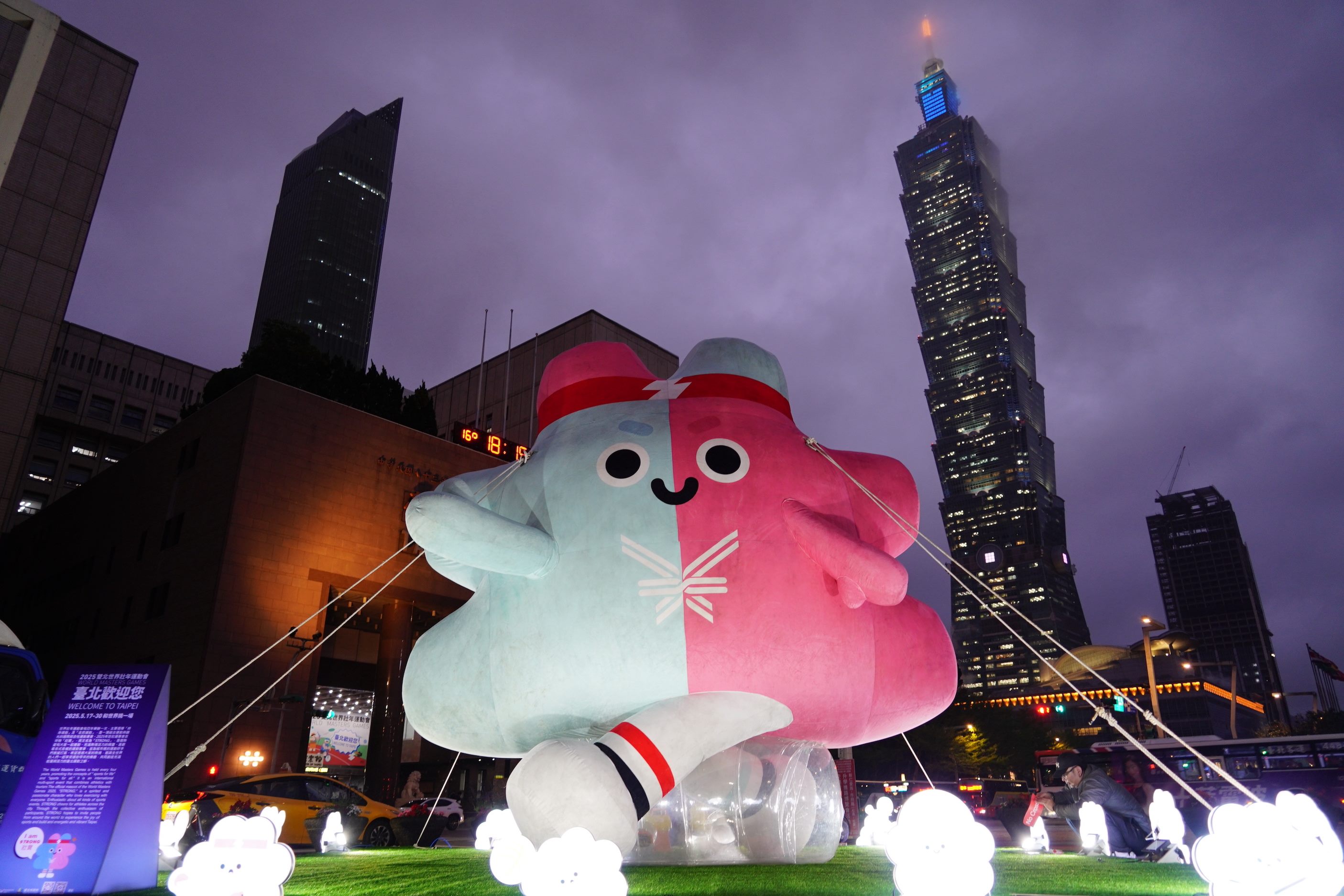 台北市信義區夜晚景象，一個巨大的粉藍雙色卡通氣球裝置坐落在市府前廣場草地上，背後可見台北101大樓燈光閃爍，天色昏暗有雲。地面有數個小型白色燈飾與一名工作人員正在調整裝置。