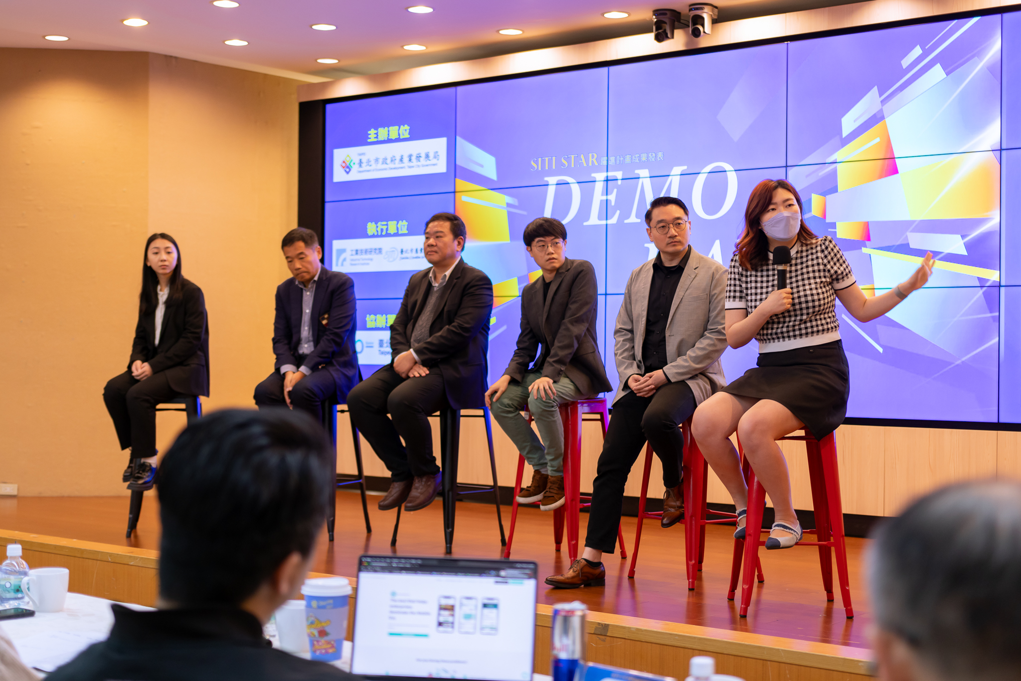 六位講者坐在舞台上高腳椅上參與 DEMO DAY 座談會，一名女性講者手持麥克風發言，背景為大螢幕顯示活動標題與主辦單位資訊，台下觀眾聚精會神聆聽。