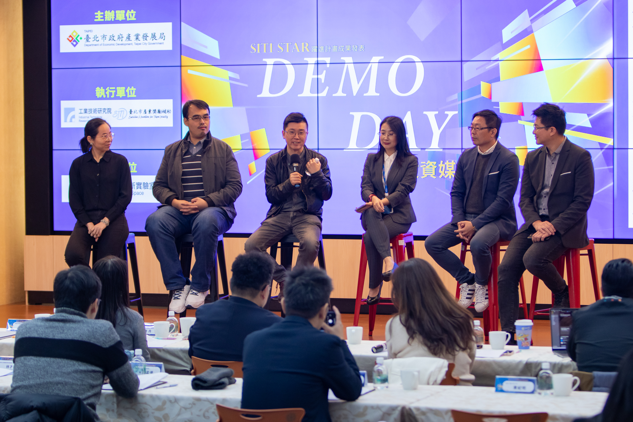 六位講者坐在舞台高腳椅上參與 DEMO DAY 座談會，中間一位男性正發言並手持麥克風，背景螢幕顯示活動標題及主辦單位資訊，台下觀眾專注聆聽。