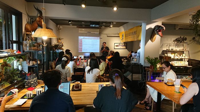 臺北創業家俱樂部活動現場，一位男性講者，微迷野林咖啡廳創辦人，呂軍逸，於投影螢幕前簡報，內容顯示表格資訊，與會者分坐多張木桌前認真聆聽並操作筆電或手機，空間布置融合植栽與動物標本，右側牆面懸掛黃色活動橫幅。