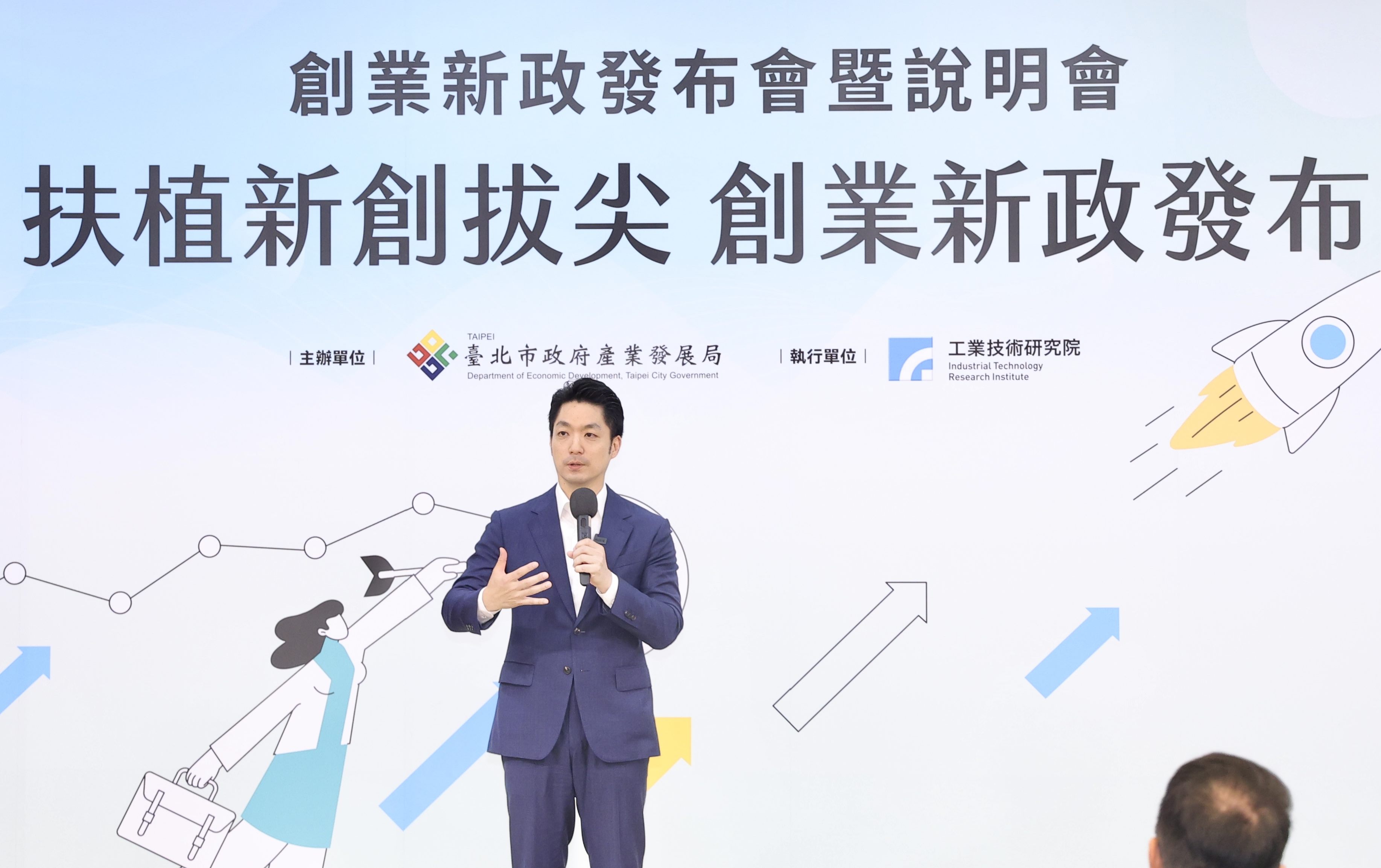 臺北市蔣萬安市長，穿著深藍色西裝，正在舞台上發表演說，背板文字為「創業新政發布會暨說明會」、「扶植新創拔尖 創業新政發布」，主辦單位為臺北市政府產業發展局，執行單位為工業技術研究院。背景設計有火箭升空、箭頭上升圖示與女性商務人物插畫，象徵創新與成長。