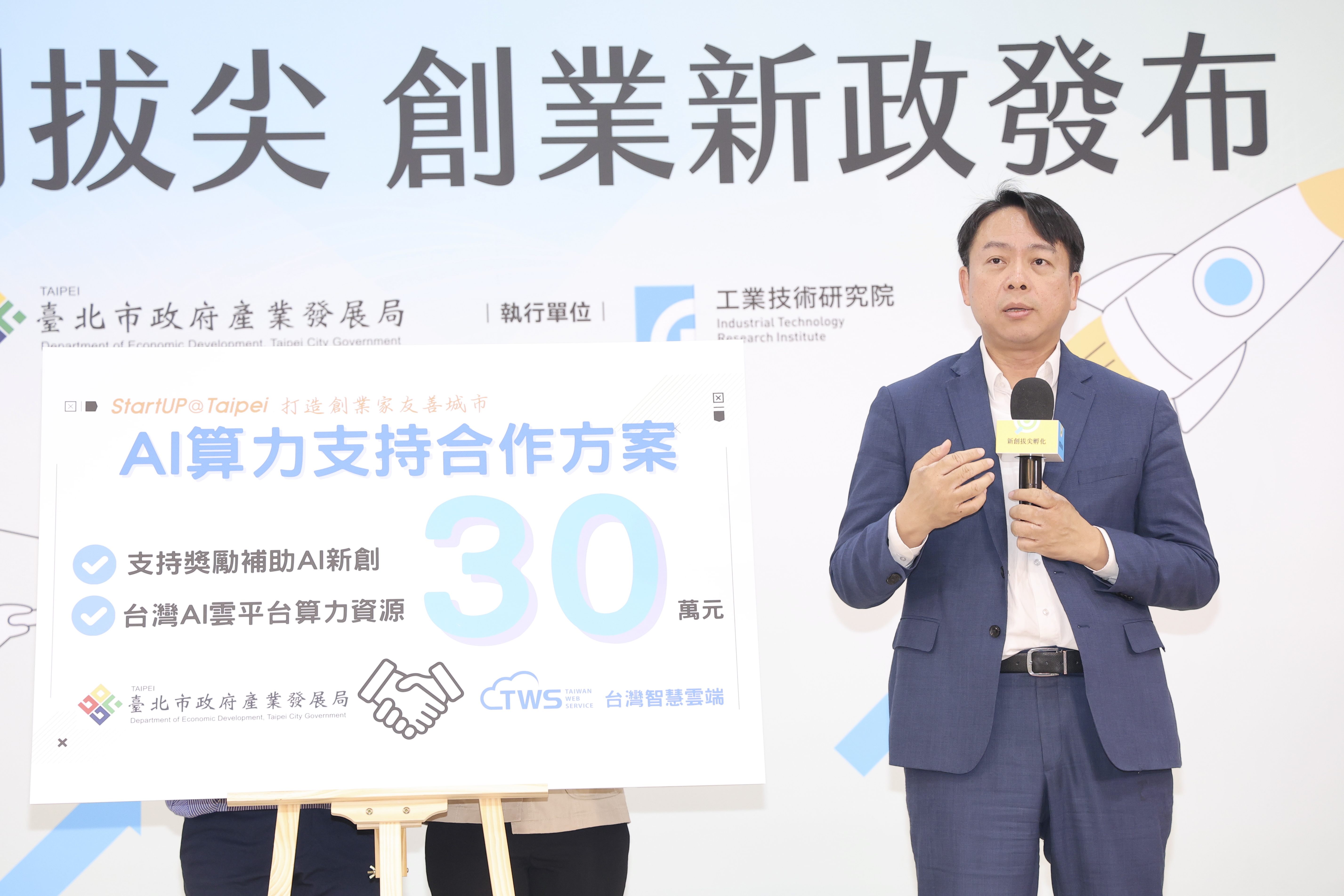 一位穿著藍色西裝的男性在創業新政發布會上發表演說，手持麥克風，站在介紹「AI算力支持合作方案」的展板旁，內容提及提供30萬元支持AI新創、台灣AI雲平台算力資源，背景為臺北市政府產業發展局與工業技術研究院的聯名標誌。